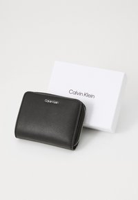 Svart läderplånbok med slät yta och silverfärgad dragkedja; pryds av en silverfärgad Calvin Klein-logga. Levereras med en vit låda.
