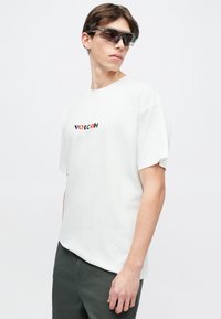 Λευκό βαμβακερό T-shirt με κοντά μανίκια και στρογγυλή λαιμόκοψη, με μαύρο και κόκκινο λογότυπο "VOLCOM" στο μπροστινό μέρος. Φορεμένο με σκούρο πράσινο παντελόνι.