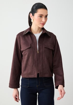 Femme aux cheveux foncés attachés en queue de cheval basse, portant des boucles d'oreilles créoles dorées, une veste marron zippée avec des poches à rabat et un jean bleu foncé devant un fond uni.