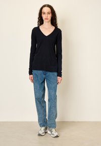 Tumesiniste varrukatega V-kaelusega pluus, mis on kombineeritud lahtiste siniste teksapükstega ja kahvatute toonidega spordijalanõudega. Sile kangas ja mugav, vaba lõige.