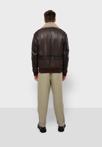 Chaqueta de cuero marrón con un cuello de borrego, con una textura suave y sutiles detalles de costura. Combinada con pantalones beige claros y zapatos negros.
