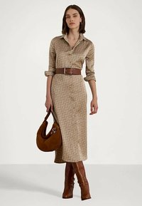 Žena v béžových vzorovaných midi šatách na gombíky s vyrolovanými rukávmi, hnedým opaskom, hnedými koženými čižmami a držiaca hnedú nubukovú kabelku.