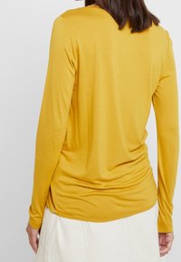 Vrouw in een mosterdgeel shirt met lange mouwen en een witte rok, gezien van achteren tegen een effen witte achtergrond.