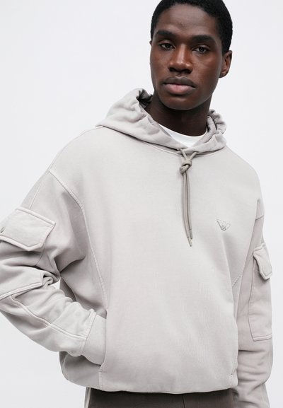 Bărbat îmbrăcat cu un hanorac gri deschis cu glugă, având buzunare mari pe mâneci și un logo mic pe piept, stând în fața unui fundal alb.