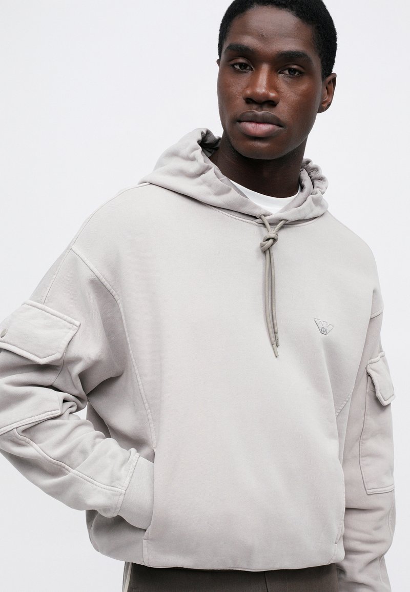 Man wearing light gray hooded sweatshirt with large pockets on sleeves and a small logo on the chest, standing against a white background.