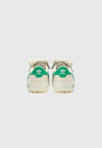 Zaļi un krēmkrāsas Adidas sporta apavi ar teksturētu režģveida virsmu, mīkstu apkakli un plakaniem šņorēm, ar izteiktu logotipu uz papēža un valkāšanas daļas.