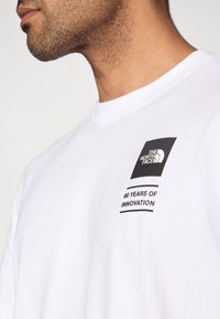 Λευκό βαμβακερό μπλουζάκι με μαύρο γραφικό που φέρει το λογότυπο "THE NORTH FACE" και το κείμενο "60 ΧΡΟΝΙΑ ΙΝΟΒΑΤΙΚΟΤΗΤΑΣ" στο αριστερό στήθος.