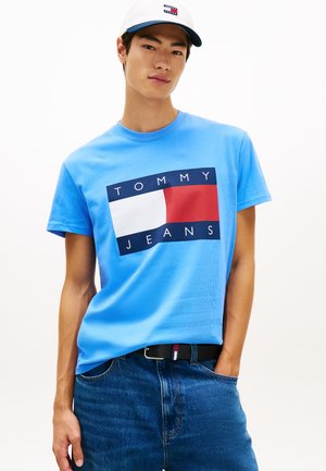 Młody mężczyzna w białej czapce, niebieskiej koszulce Tommy Jeans z logo, niebieskich jeansach i czarnym pasku, pozujący z jedną ręką w kieszeni.