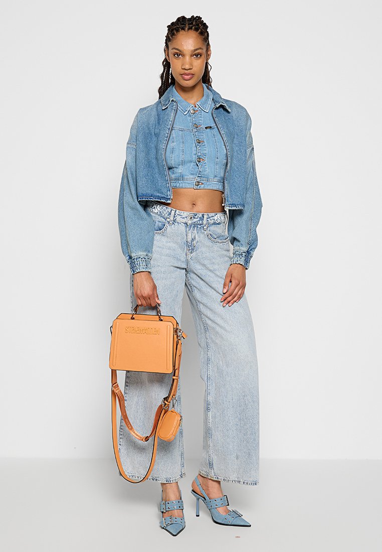 Donna in giacca di jeans corta, pantaloni larghi a vita alta, tacchi blu con borchie e che tiene una borsa arancione con tracolla.