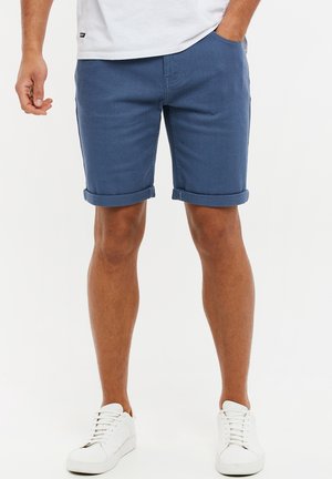 Mand iført opkrogede blå shorts, hvide sneakers og en hvid t-shirt med den ene hånd i lommen, stående på en hvid baggrund.
