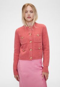 Rózsaszín kötött kardigán lekerekített gallérral, aranyszínű gombokkal, két elülső zsebbel és bordázott mandzsettákkal és szegéllyel. Simogató tapintású, testhezálló kialakítás.