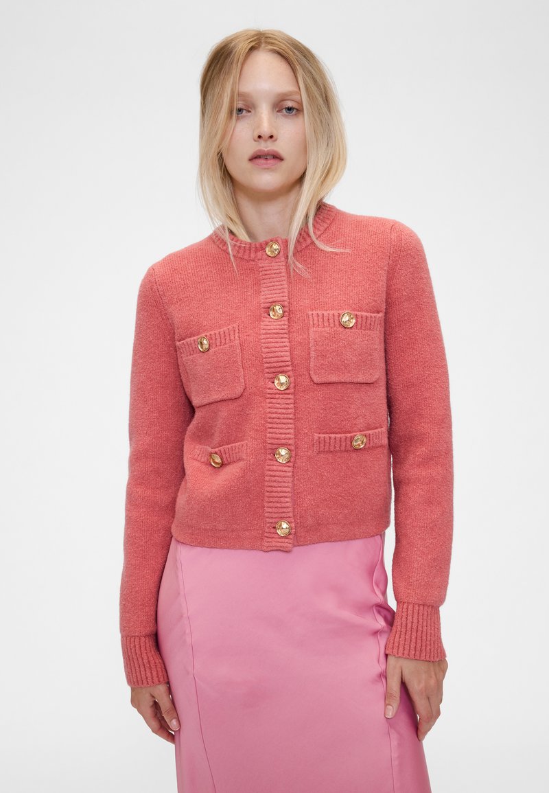 Rózsaszín kötött kardigán lekerekített gallérral, aranyszínű gombokkal, két elülső zsebbel és bordázott mandzsettákkal és szegéllyel. Simogató tapintású, testhezálló kialakítás.