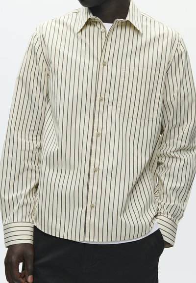 Chemise boutonnée à rayures crème avec lignes horizontales et verticales, une poche unique et des poignets à boutons. Tissu en coton léger.