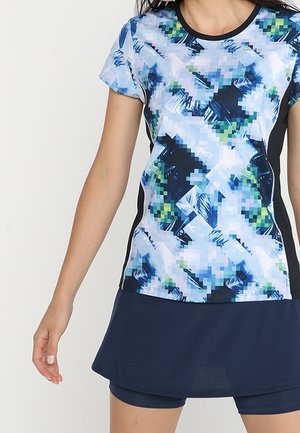 Kobieta ubrana w sportową koszulkę z krótkim rękawem o wzorze pikselowym w kolorach niebieskim, białym i zielonym, połączoną z granatową spódnicą i szortami.