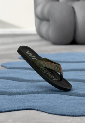 Czarna sandałka typu klapek z oliwkowozielonymi, plecionymi paskami, oznaczona marką "Wrangler", spoczywająca na niebieskiej teksturowanej macie na rozmytym, szarym tle.