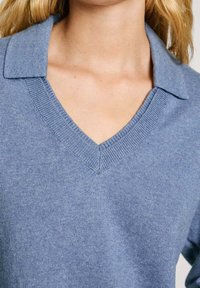 Gros plan d'une personne portant un pull en tricot bleu avec un col en V et un détail de col replié.