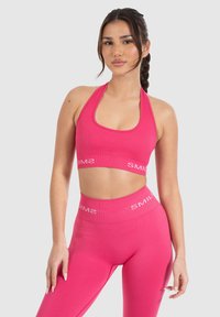 Mujer con el cabello trenzado que lleva un sujetador deportivo tipo halter rosa brillante a juego y leggings de cintura alta con la marca "SMIL" en blanco.