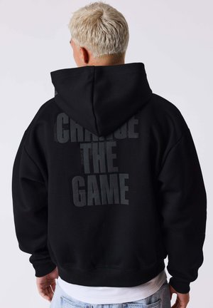 Personne aux cheveux blonds courts portant un sweat à capuche noir avec le texte « CHANGE THE GAME » au dos, associé à un jean bleu clair.