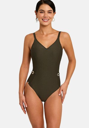 Lachende vrouw in een olijfgroene eendelige badpak met dunne bandjes en decoratieve zijgespen, staand tegen een effen achtergrond.