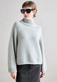 Světle modrý pletený svetr s vysokým žebrovaným límcem, spadlými rameny a dlouhými rukávy, v kombinaci s tmavou sukní. Viditelné detaily texturované tkaniny.
