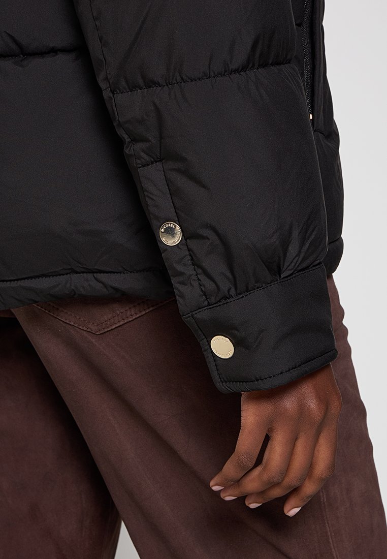 Chaqueta puffer negra con un acabado texturizado. Presenta un primer plano del puño con botones de hardware dorados y un diseño ligeramente acolchado.