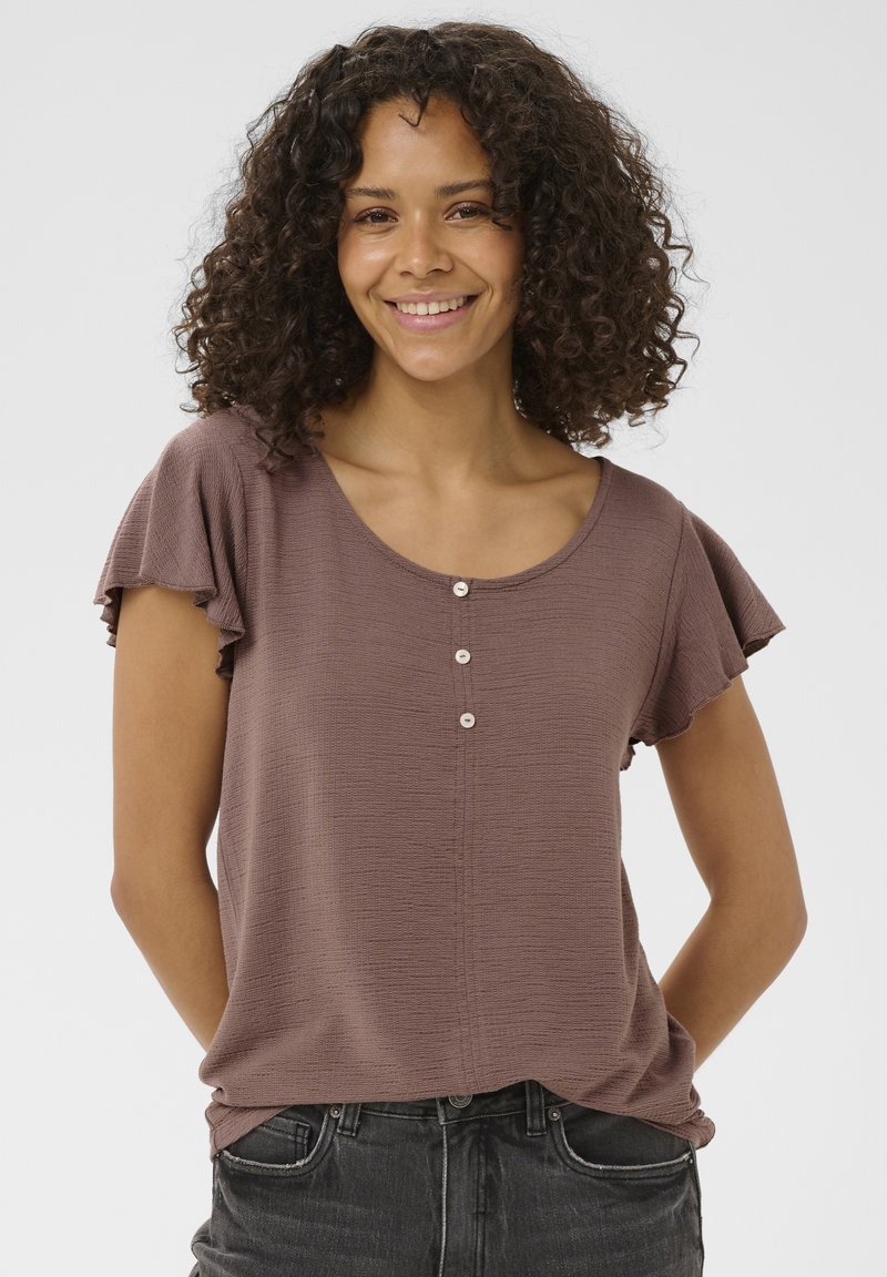 Femme souriante aux cheveux bouclés portant un haut mauve à manches courtes avec trois boutons et un jean noir délavé, debout devant un fond uni.