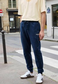 Pantaloni de trening bleu marin cu talie elastică și broderie aurie, asociați cu adidași albi cu dungi negre și o cămașă crem cu mânecă scurtă.