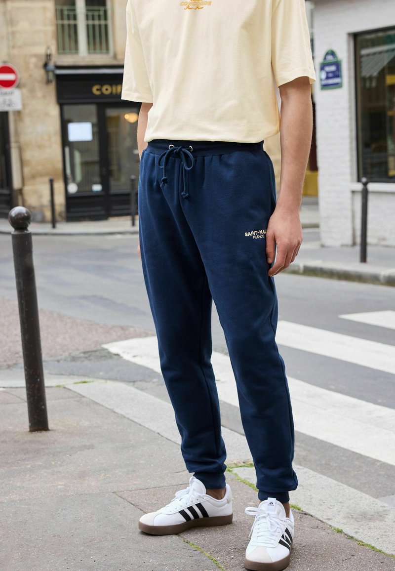 Pantaloni de trening bleu marin cu talie elastică și broderie aurie, asociați cu adidași albi cu dungi negre și o cămașă crem cu mânecă scurtă.