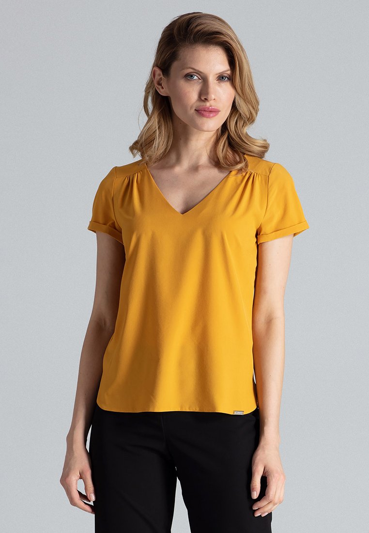 Gelbe V-Ausschnitt-Bluse mit kurzen Ärmeln, aus weichem Stoff und subtilen Raffungselementen an den Schultern. Kombiniert mit schwarzen Hosen.