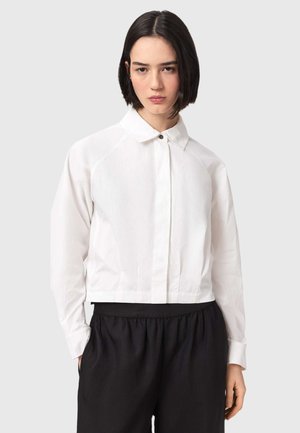 Vrouw met kort donker haar, gekleed in een wit overhemd met lange mouwen en knopen, en losse zwarte broek, staand tegen een effen lichte achtergrond.