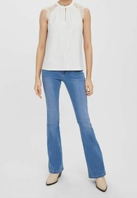 Top bianco senza maniche con dettagli in pizzo sulle spalle e apertura a chiave sul collo; abbinato a jeans svasati blu e scarpe col tacco beige.