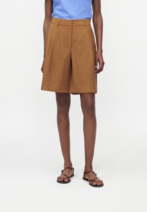 Frau trägt braune knielange Shorts, ein hellblaues Top und braune Sandalen mit Riemchen, steht vor einem schlichten weißen Hintergrund.