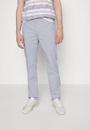 Persona que lleva pantalones casuales gris claro, zapatillas blancas y una camiseta sin mangas a rayas, de pie frente a un fondo blanco liso.