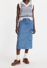 Pletená vesta s modro-krémovými vzory přes džínsovou midi sukni s postranními kapsami, doplněná hnědými šněrovacími botami a krémovými ponožkami.