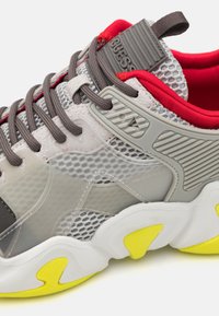 Zapatilla de malla gris con acentos rojos, suela gris y amarilla, detalles texturizados y una etiqueta de marca en la lengüeta. Los cordones en marrón oscuro completan el diseño.