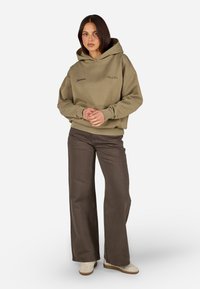 Olivgrüner Kapuzenpullover mit Fronttasche; hat schwarze Logos. Kombiniert mit braunen weiten Hosen und beigen Sneakers.