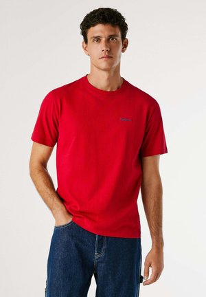 Camiseta de algodón roja con cuello redondo, mangas cortas y un logo bordado azul en el pecho. Combinada con pantalones de mezclilla azules.