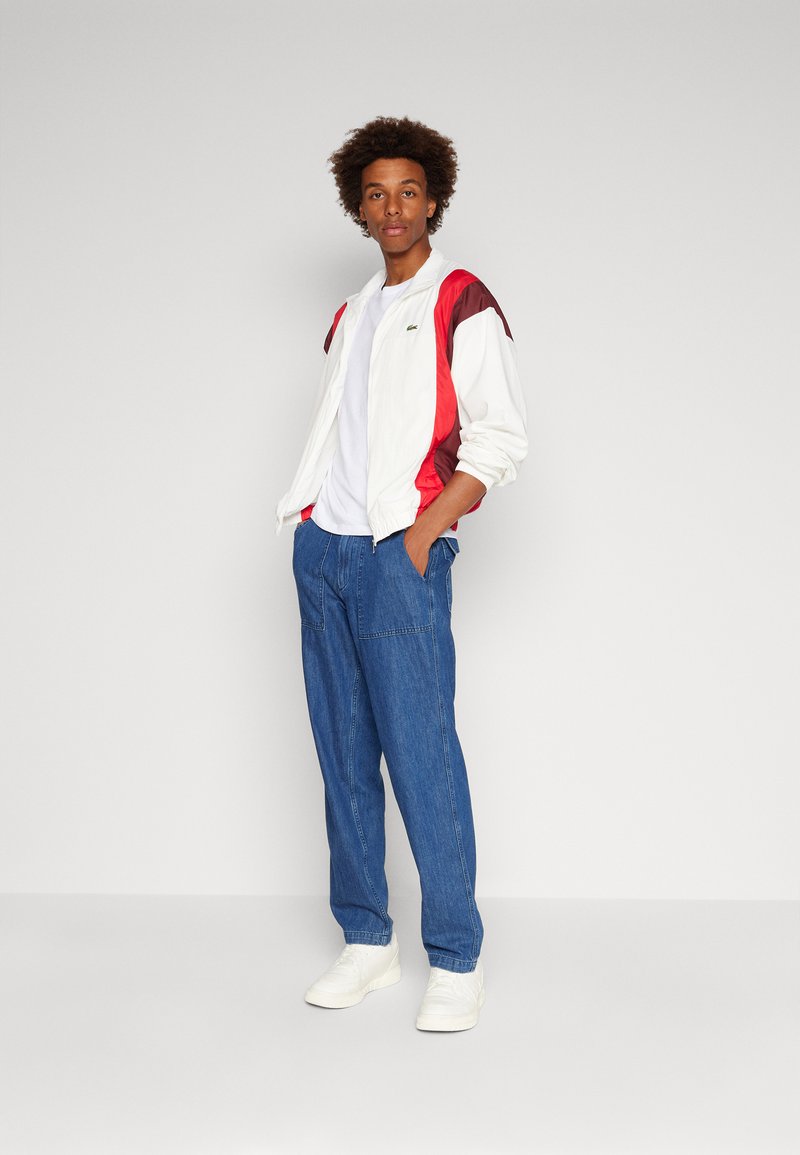 Veste sportive blanche et rouge, pantalon en jean bleu, t-shirt blanc et baskets blanches. Le modèle se tient les mains dans les poches, avec les cheveux courts et bouclés.