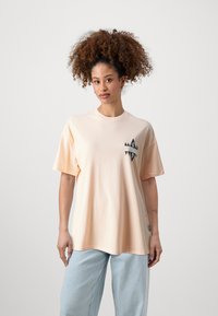 Peach-colored oversized t-shirt with short sleeves, featuring a black geometric logo on the chest and a crew neckline.