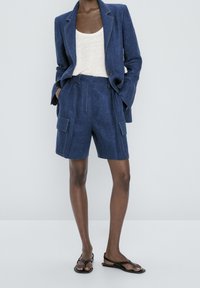 Personne portant un blazer bleu marine et un short cargo assorti avec une chemise blanche et des sandales noires, les mains dans les poches, debout devant un fond uni.