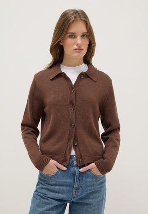 Jeune femme portant un cardigan marron boutonné sur un top blanc, les mains dans les poches d’un jean bleu clair, sur un fond uni.