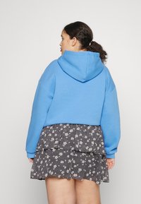 Casaco de algodão azul com capuz e mangas longas, combinado com uma saia preta floral em camadas com detalhes brancos.