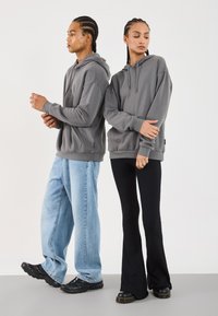Le felpe grigie presentano un tessuto morbido, tasche a marsupio e cappucci con lacci. Un modello indossa jeans baggy azzurri; l'altro indossa pantaloni svasati neri.