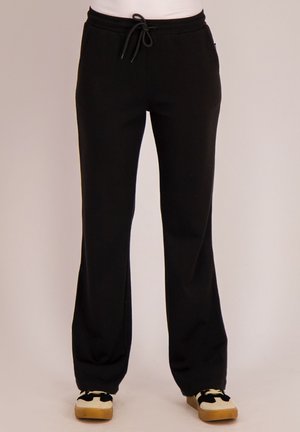 Zwarte broek met trekkoord gedragen door een persoon die staat, gecombineerd met witte en zwarte sneakers met gumzolen.