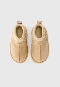 Deux pantoufles beige en suede doux, avec un col en tricot côtelé et une petite languette à l'arrière pour un enfilage facile.