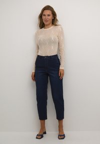 Durchscheinendes, beiges Langarm-Oberteil mit Blumenmuster, kombiniert mit hoch taillierten dunkelblauen Jeans. Offene, navyblaue Absätze runden das Outfit ab.