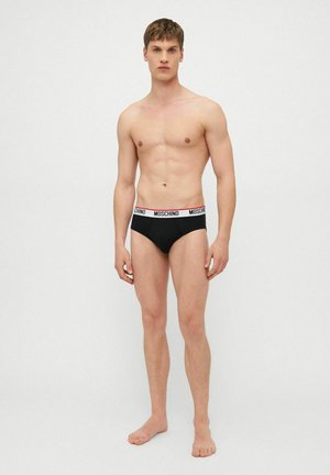 Giovane uomo in piedi a piedi nudi che indossa slip neri con fascia elastica bianca con il logo "Moschino", su sfondo chiaro e uniforme.