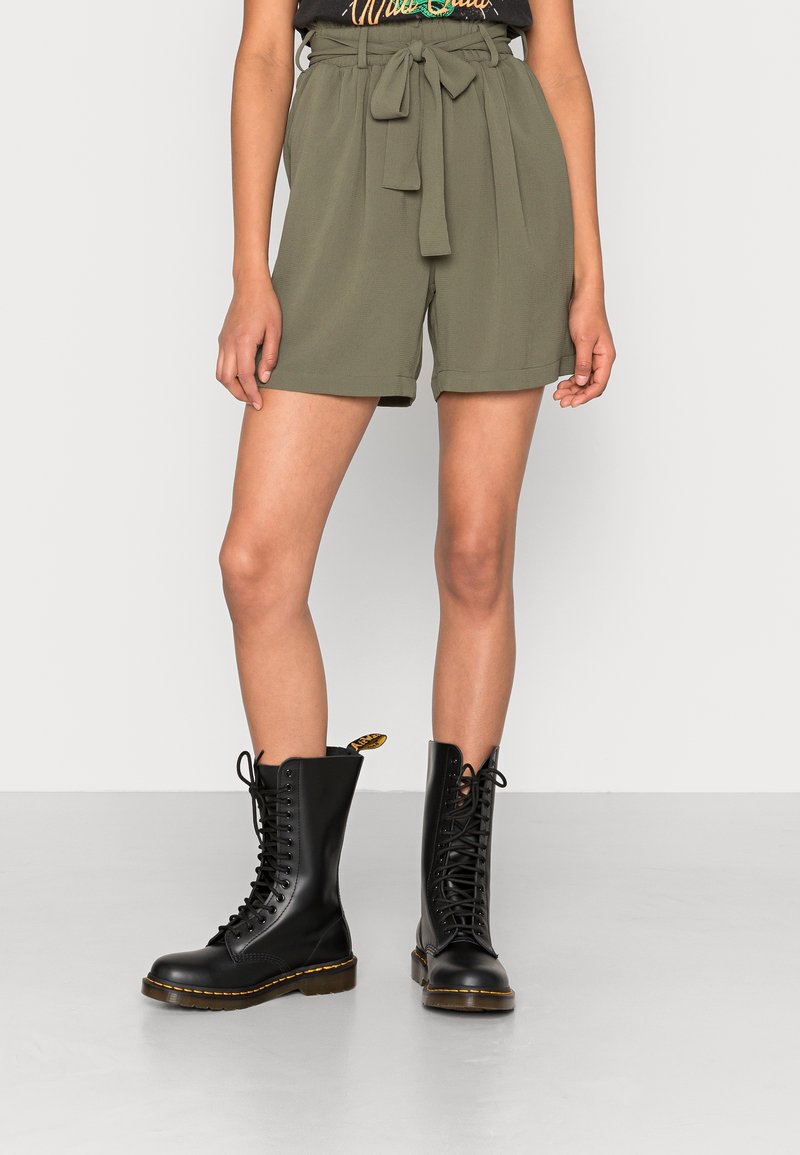 Pantaloni corti alta vita verde oliva con dettaglio della cintura a fiocco, abbinati a stivali da combattimento neri con lacci, caratterizzati da una suola chunky e cuciture gialle.