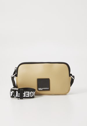 Kremfarget skinn crossbody-veske med svarte detaljer, glidelåslukking, rektangulær form og logo-patch. Har justerbar stropp med tekstmønster.