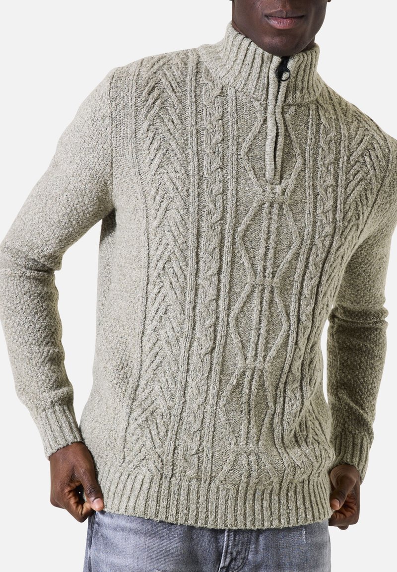 Lichtgrijze gebreide trui met een hoge kraag, gekenmerkt door een getextureerd kabelbreipatroon en ribbels aan de mouwen en onderrand. Voorzijde met ritssluiting.